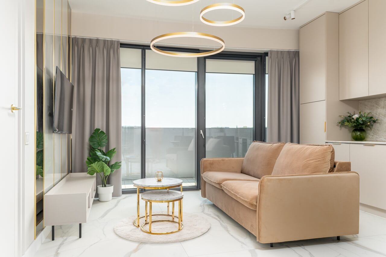 Salon z beżową sofą, marmurowymi stolikami i dużymi przeszkleniami prowadzącymi na balkon