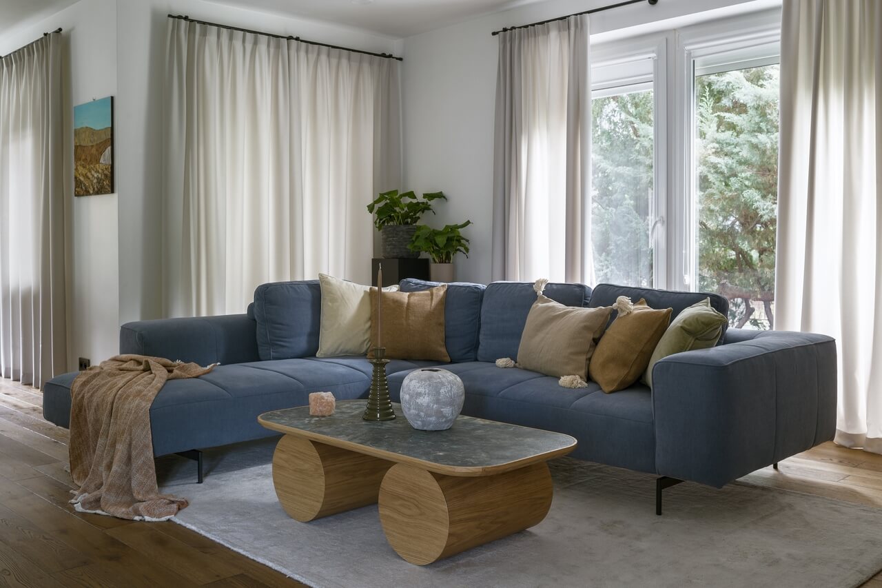 Niebieska sofa narożna w salonie, stolik kawowy z drewnianymi detalami, zasłony