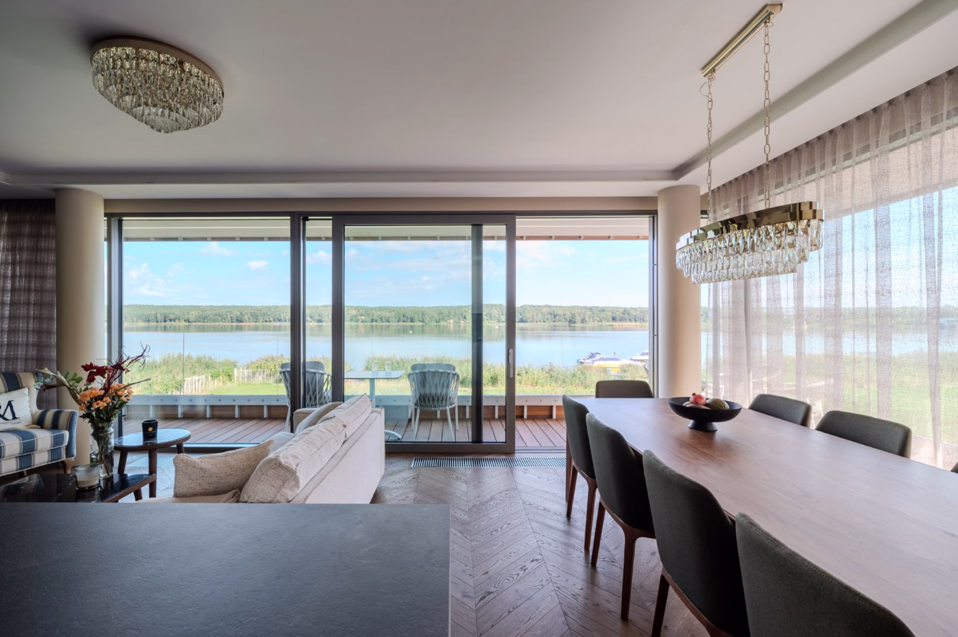 A modern living room with a view of the lake, elegant chandeliers, wooden flooring.