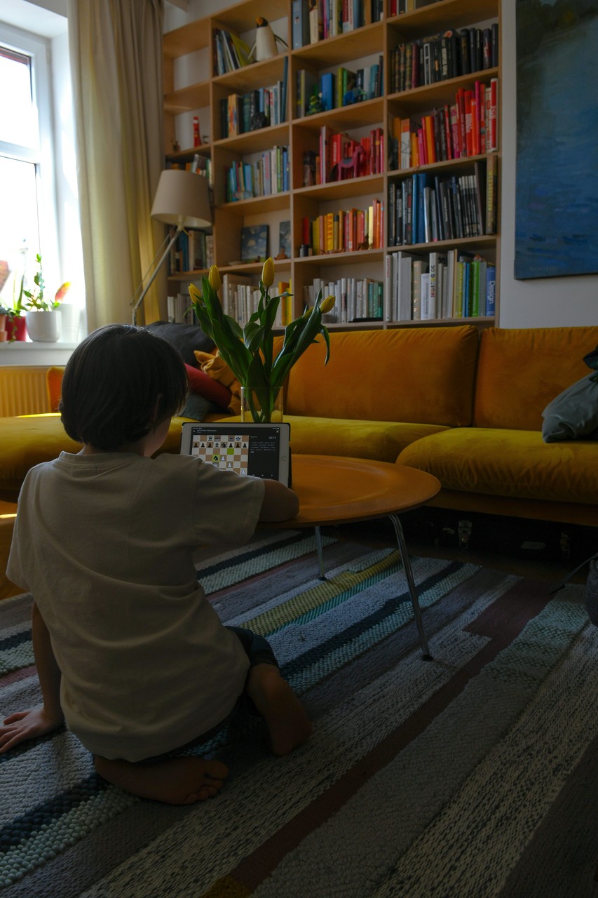 Dziecko gra w szachy na tablecie w przytulnym salonie z biblioteczką i żółtą kanapą.