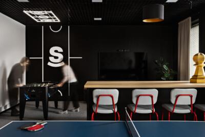 Nowoczesna przestrzeń biurowa z designerskimi krzesłami, telewizorem i stołem do ping-ponga. Industrialny sufit i ściana z logiem dodają charakteru wnętrzu.