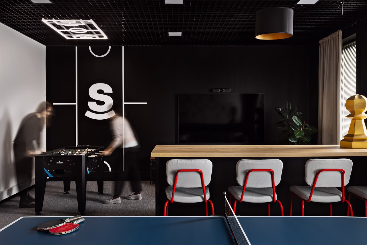 Nowoczesna przestrzeń biurowa z designerskimi krzesłami, telewizorem i stołem do ping-ponga. Industrialny sufit i ściana z logiem dodają charakteru wnętrzu.