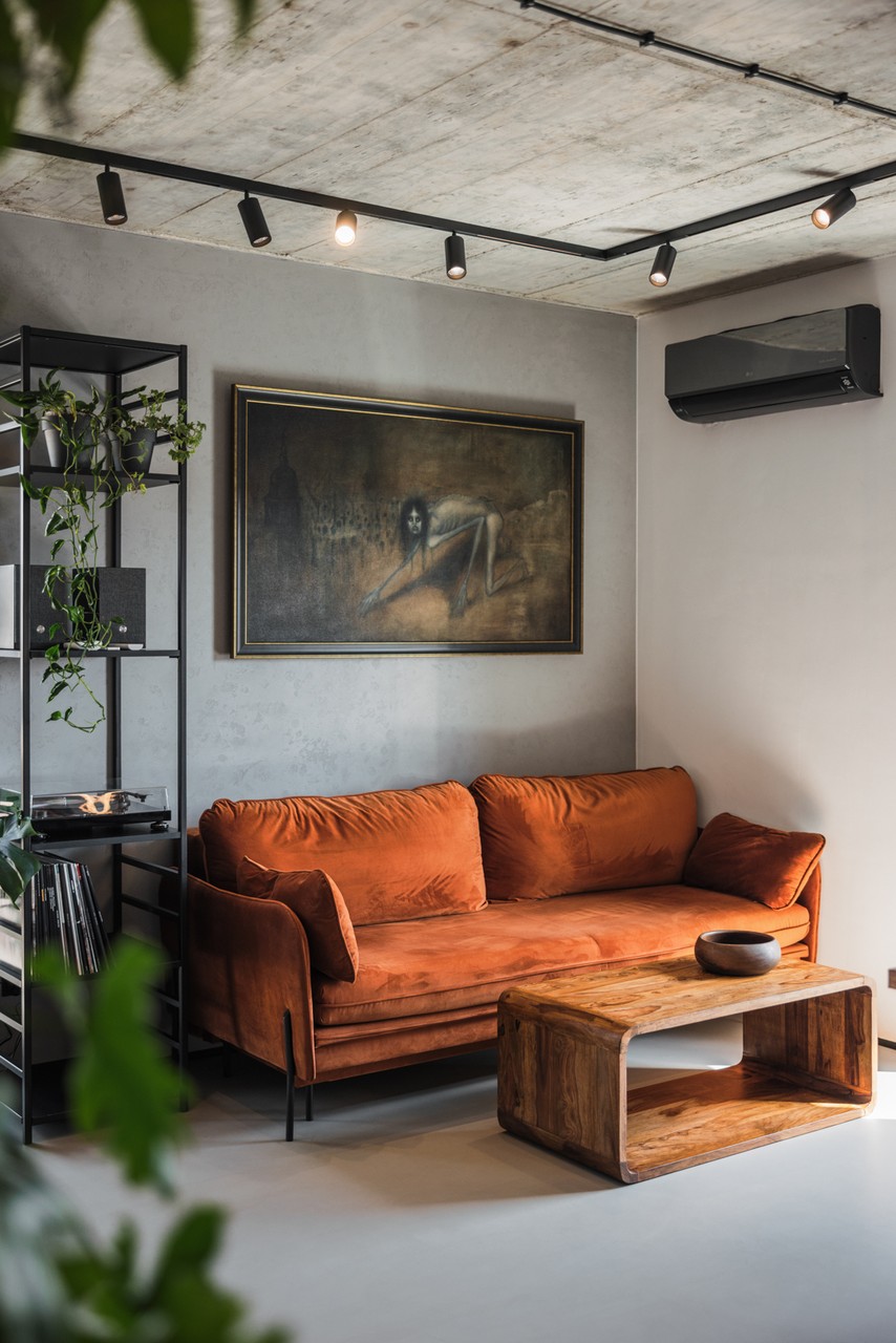 Industrialny salon z sofą w odcieniach rudości, obrazem i zielenią.