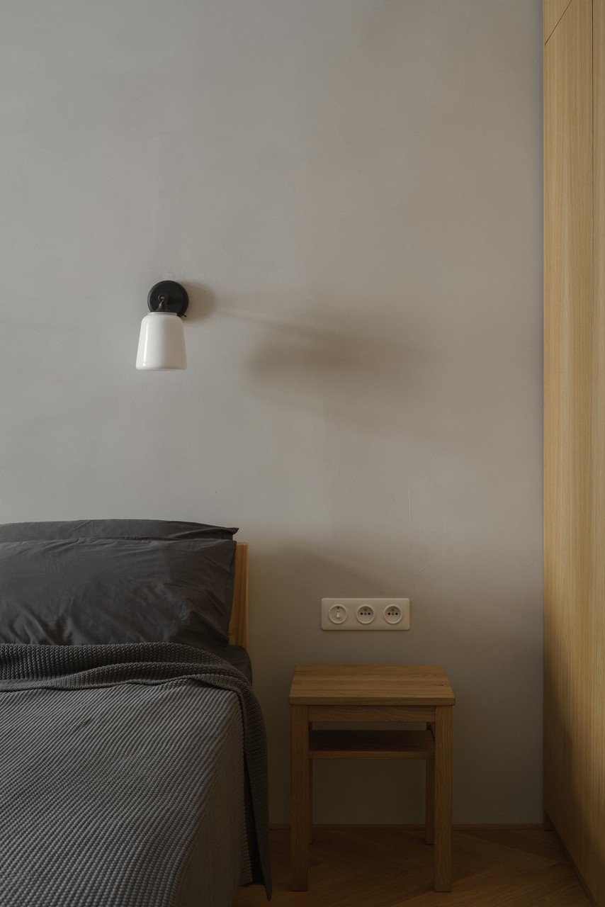 Minimalistyczna sypialnia z lampą ścienną i drewnianym stolikiem.