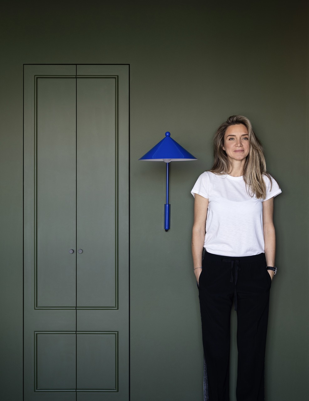 Kobieta przy oliwkowej ścianie z niebieską designerską lampą.