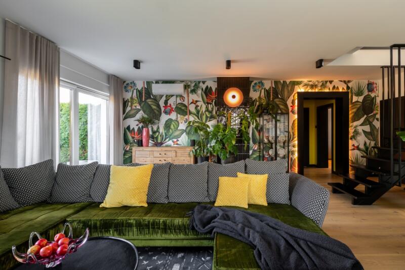 Salon z zieloną sofą, botaniczną tapetą i żółtymi poduszkami.