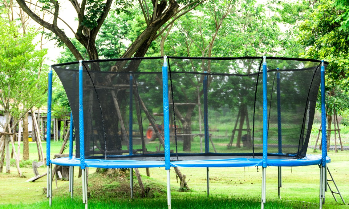 Trampolina ogrodowa z siatką pośród zieleni i drzew.