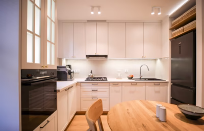 Jasna kuchnia z białymi szafkami, drewnianym stołem i czarną lodówką.