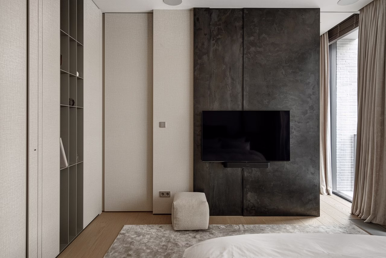 Minimalistyczna sypialnia z betonową ścianą, telewizorem i beżowymi zasłonami.