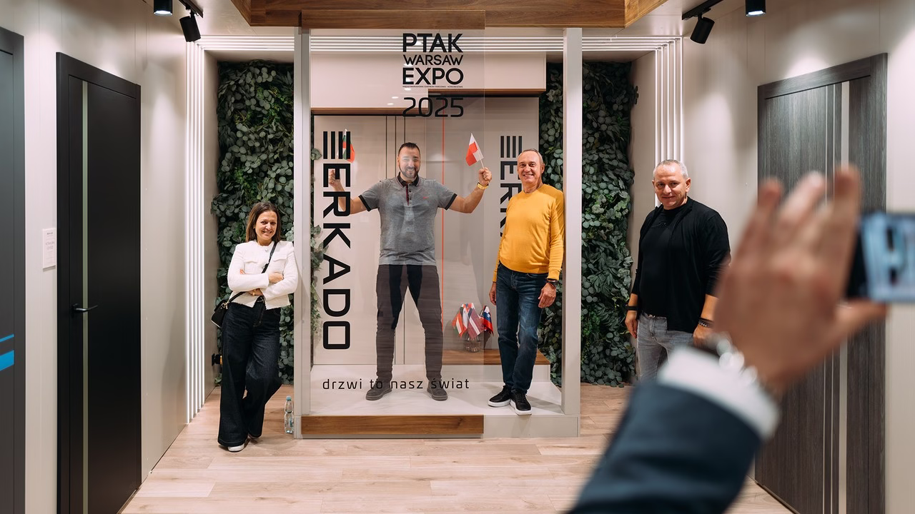 Ludzie pozują przed stoiskiem Erkado na tle zielonych ścian.