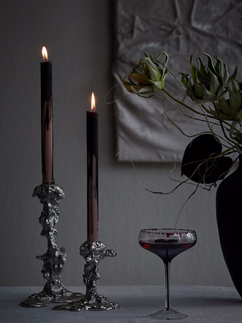 Czarne świece na fantazyjnych świecznikach, kieliszek i kwiaty w wazonie.