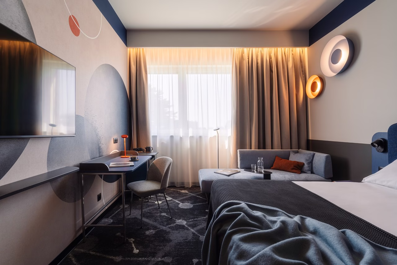 Nowoczesny pokój hotelowy z designerskim oświetleniem, biurkiem i szarą sofą.