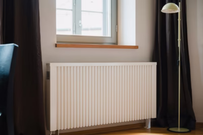 Biały grzejnik pod oknem, obok kremowa lampa, ciemne zasłony.