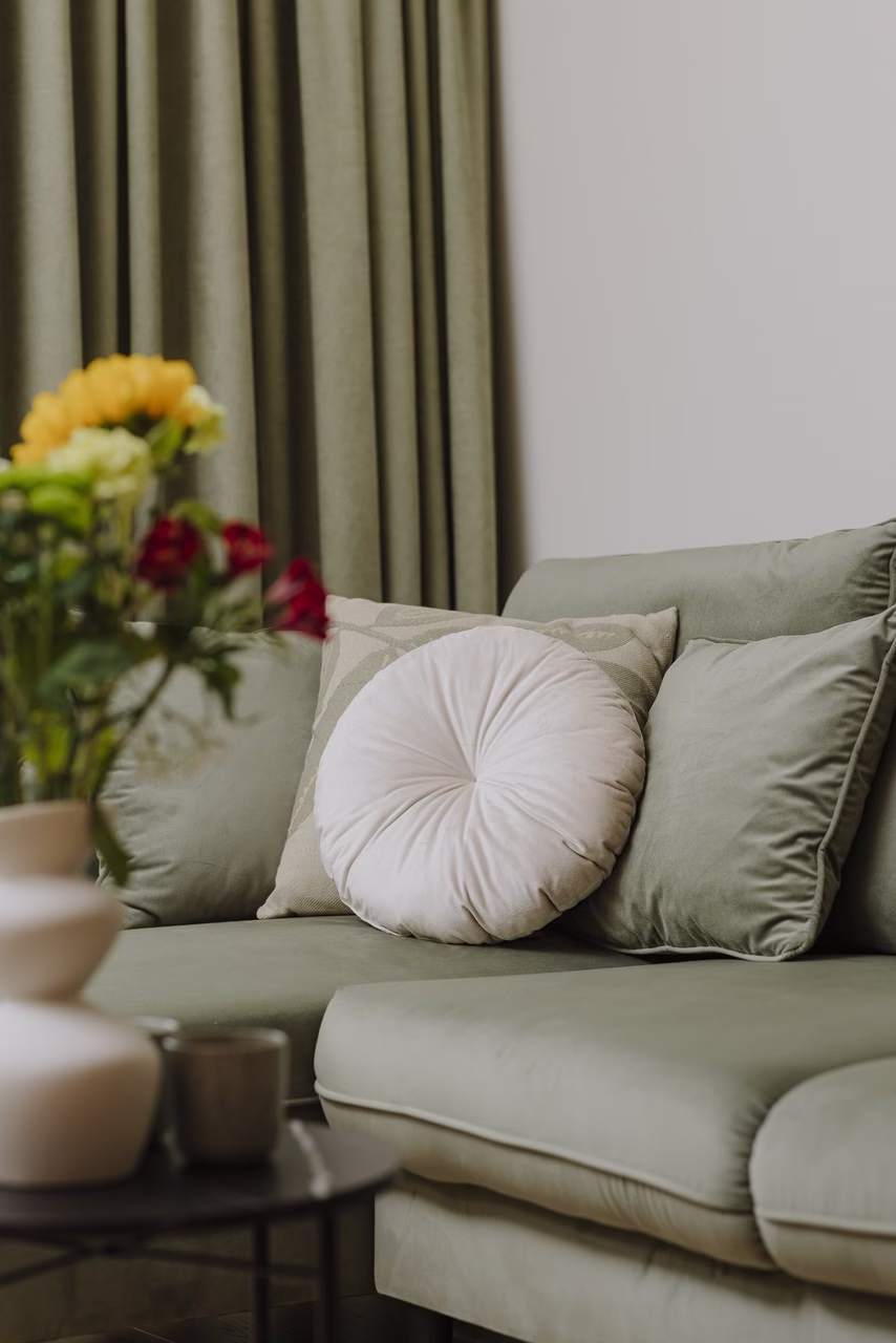 Zielona sofa z okrągłą poduszką, kwiaty w wazonie na stoliku.