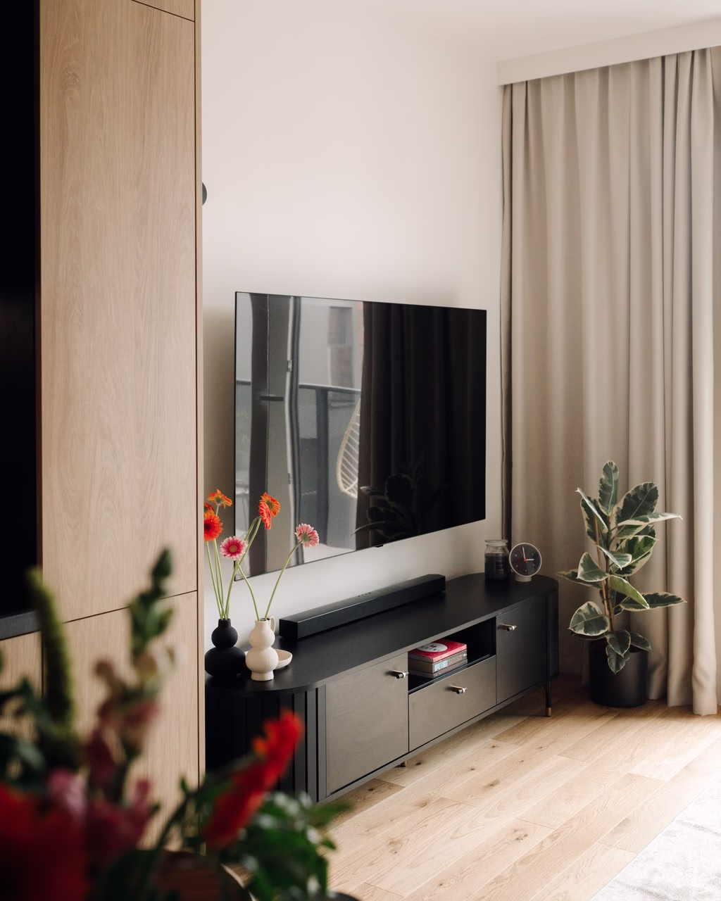 Nowoczesny salon z telewizorem, ciemnymi meblami, jasnymi zasłonami i rośliną.
