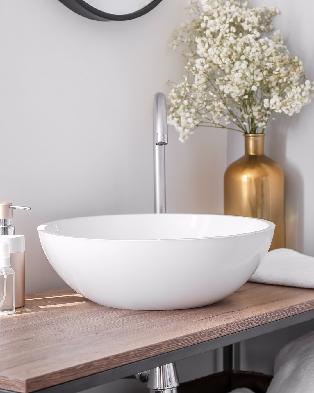 Biała umywalka na drewnianym blacie z złotym wazonem i kwiatami.