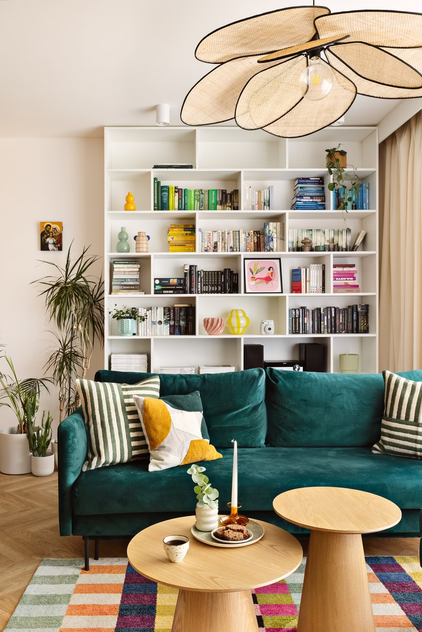 Zielona welurowa sofa, drewniane stoliki, jasna półka i kolorowy dywan.