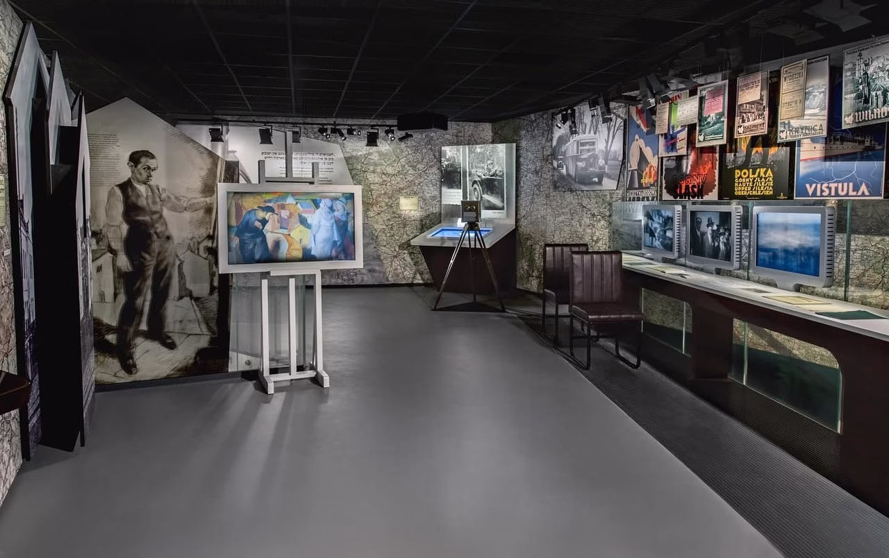 Muzealne wnętrze z mapami, plakatami, starymi zdjęciami i ekranami komputerowymi.