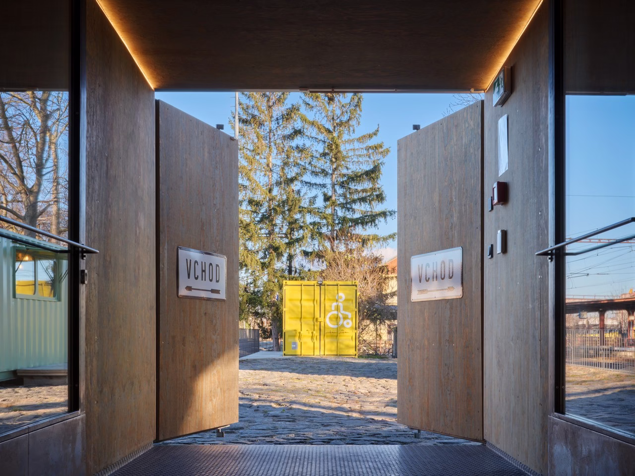 Nowoczesne wejście z drewnianymi drzwiami, widoczny żółty kontener na zewnątrz.
