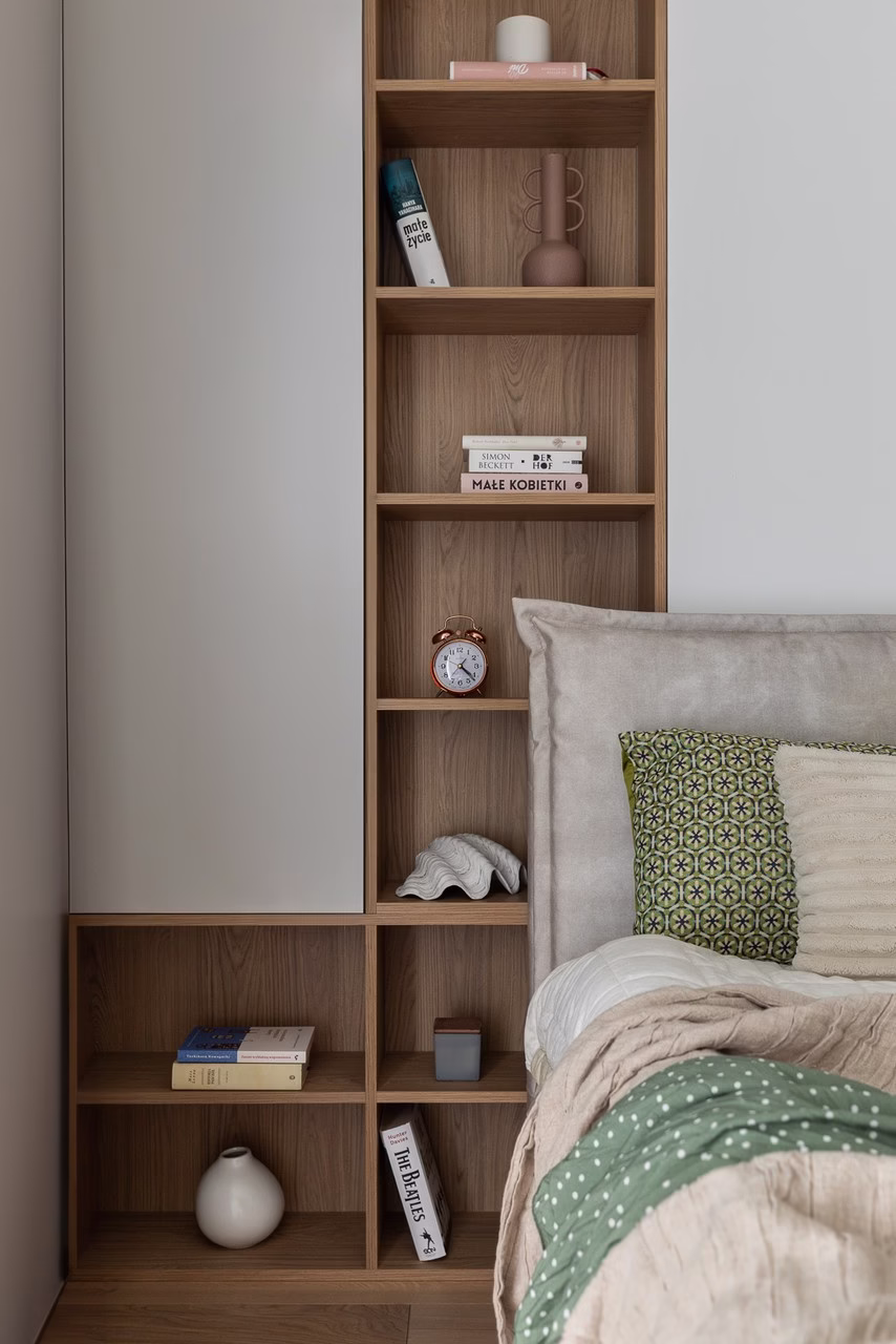 Drewniany regał w jasnej sypialni z książkami i wzorzystą poduszką.
