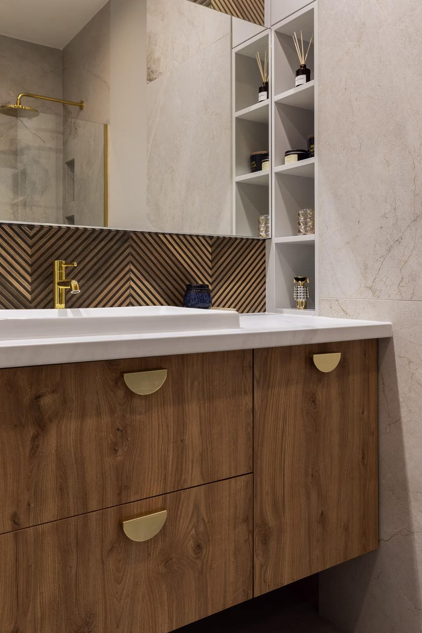 Elegancka łazienka z drewnianą szafką, złotymi uchwytami i lustrzanym panelem.