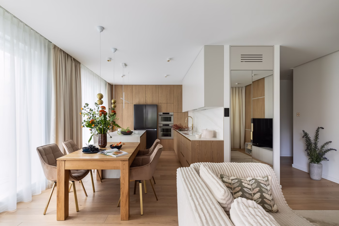 Nowoczesna kuchnia z drewnianymi meblami, jasnymi zasłonami i kwiatową dekoracją.
