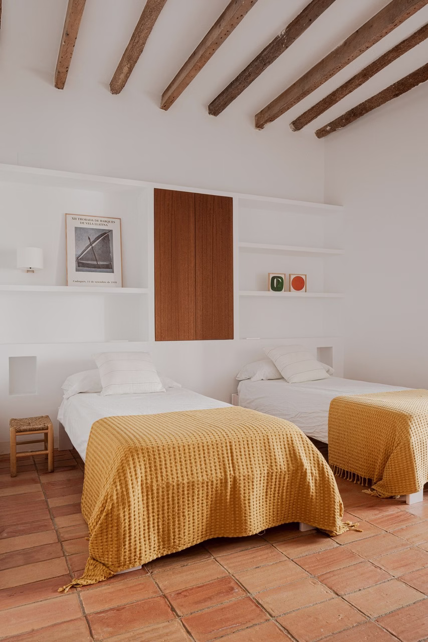 Dwie łóżka z musztardowymi narzutami, białe półki, drewniane belki.